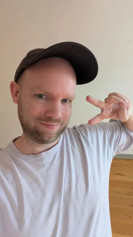 A man in a dark baseball cap and a pale t-shirt taking a selfie indoors, holding up two fingers