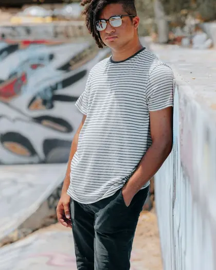A young man with dreadlocks and glasses in a striped t-shirt, standing against a graffitied wall