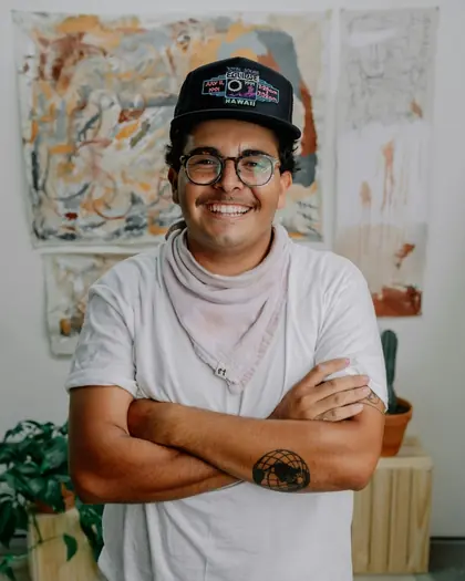 A smiling man in a cap and white t-shirt with his arms folded, standing indoors in front of hanging artwork